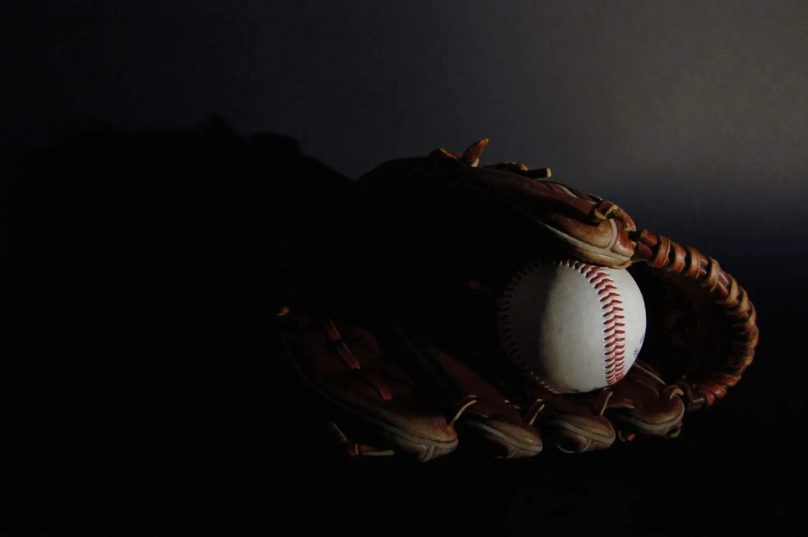 A baseball and mitt in the dark with light shining on it.